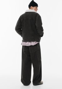 Giacca in velluto a coste nera con un colletto in pelliccia sintetica bianca, indossata sopra una camicia a quadri rosa, abbinata a pantaloni neri larghi.