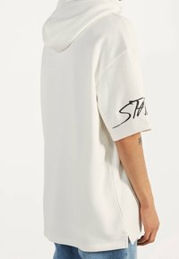 Vit oversized hoodie i mjukt material med huva, korta ärmar och sidofrackar. Har fet svart text på vänster ärm.