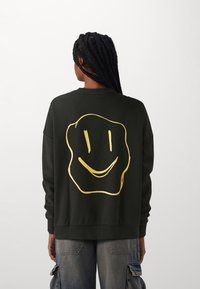 Černá mikina s velkým zlatým potiskem usměvavé tváře na zadní straně. Má pohodlný střih a dlouhé rukávy s manžetami.