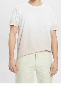 T-shirt bianca in cotone con un morbido gradiente che sfuma verso un rosa chiaro sull'orlo, maniche corte e colletto a girocollo; abbinata a pantaloni beige chiaro.