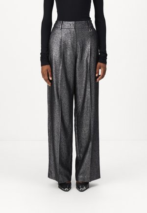 Personne portant un pantalon argenté métallique taille haute et jambes larges, un haut noir à manches longues et des chaussures noires pointues, debout devant un fond uni.