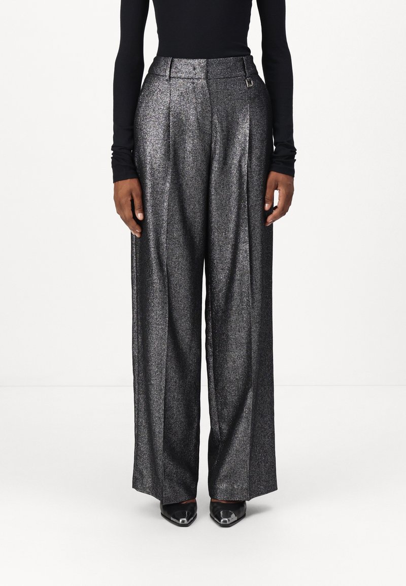 Personne portant un pantalon argenté métallique taille haute et jambes larges, un haut noir à manches longues et des chaussures noires pointues, debout devant un fond uni.