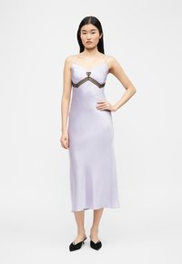 Γυναίκα που φοράει μίντι φόρεμα από ανοιχτό μωβ σατέν χωρίς μανίκια, με λεπτές ράντες και μαύρη δαντέλα κάτω από το στήθος, στέκεται με το ένα χέρι στη μέση.