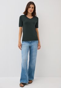 T-shirt vert foncé à manches courtes en V associé à un jean bleu clair évasé et des sandales noires avec des accents métalliques.