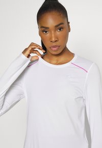 Valge pika varrukaga spordipusakas roosa aktsendiga õlgadel ja väikese Puma logoga, ümara kaelusega ja sileda tekstuuriga.