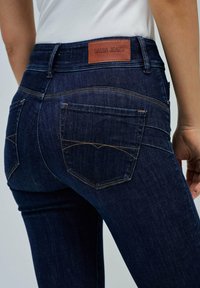 Jeans de mezclilla azul oscuro con costuras naranjas, que cuentan con una etiqueta de cuero en la cintura y dos bolsillos traseros con un diseño sutil.