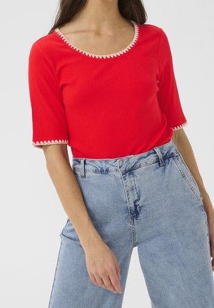 Czerwony prążkowany top z dekoltem w kształcie litery U i kremową falbanką. Połączony z jasnobeżowymi, wysokim stanem jeansami. Krótkie rękawy i gładka faktura.