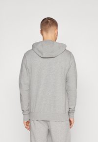 Mand med kort hår iført en ensfarvet grå hættetrøje og matchende joggingbukser, set bagfra mod en hvid baggrund.