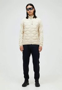 Mann med langt svart hår som har på seg svarte solbriller, beige kviltet jakke med glidelås, svarte bukser og svarte joggesko, står mot hvit bakgrunn.