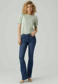 Gaiši zaļš īsām piedurknēm tops ar puffed piedurknēm, apvienots ar tumši ziliem platām biksēm un bēšiem augstpapēžu sandalēm uz neitrāla fona.