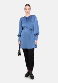 Blauwe satijnen blouse met een hoge halslijn, lange pofmouwen, een gestrikte taille en zij-splitten. Zwarte broek en schoenen complementeren de outfit.