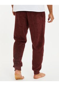 Bordeaux fleece loungebroek met een pluizige textuur, elastische tailleband en omgeslagen enkels. Geen zichtbare zakken. Voeten zijn bloot.
