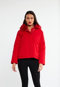 Röd puffjacka med fuskpälskrage, dragkedja och sidofickor. Har en kort design och texturerad tyg. Svarta leggings bärs under.