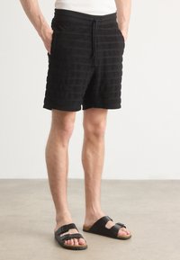 Zwarte shorts met verticale texturen strepen, elastische tailleband en trekkoord, gecombineerd met zwarte instapsandalen met een gespsluiting.
