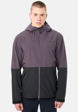 Jonge man met een tweekleurig paars-zwart capuchonjack met rits, staand tegen een effen lichte achtergrond.