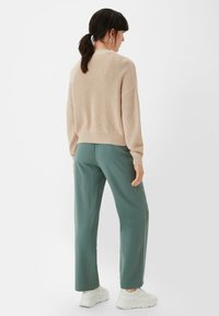 Beige gestrickter Pullover mit geripptem Saum, kombiniert mit grünen weiten Hosen. Abgerundet mit weißen Sneakern. Einfarbiger Hintergrund.