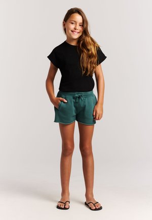 Lachend meisje met lang haar dat een zwart shirt met korte mouwen draagt, turquoise shorts met een trekkoord en zwarte slippers, staand met één hand in de zak.