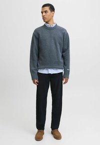 Un pull en tricot gris avec un col côtelé, porté par-dessus une chemise rayée bleue, associé à un pantalon en velours côtelé foncé et des chaussures en daim marron.
