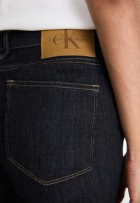 Mörk denimjeans med en slät yta, brunt lädermärke med präglad "Calvin Klein Jeans" och kontraststygn på fickorna.