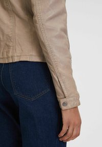 Lichtbeige leren jack met een getailleerd ontwerp, zichtbare stiksels en drukknoopdetails bij de mouwmanchet. Donkerblauwe denim jeans zichtbaar.