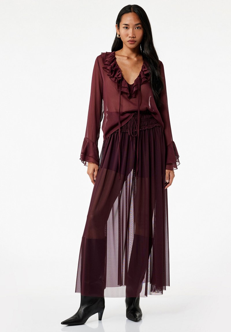 Bordeaux-gerimpelde blouse met een diepe V-hals, gecombineerd met een bijpassende doorschijnende rok. Bevat lange mouwen en een taille met trekkoord. Zwarte puntige laarzen.