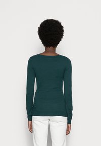 Personne aux cheveux noirs courts et bouclés, portant un haut à manches longues vert foncé et un pantalon blanc, debout face à un fond blanc uni.