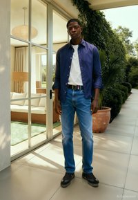 Homem vestindo uma camisa azul-marinho abotoada por cima de uma T-shirt branca, jeans azuis e tênis pretos, em pé ao ar livre perto de portas de vidro e vegetação.