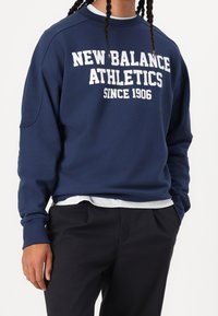 Tummansininen huppari, jossa valkoinen "New Balance Athletics Since 1906" -teksti. Valmistettu sileästä kankaasta, jossa on pyöreä kaula-aukko ja resorit hihoissa.