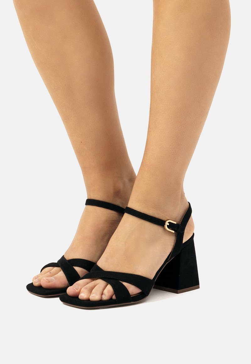 Sandali in suede neri con tacco a blocco, design a fascetta incrociata e fibbia alla caviglia. Punta aperta. Materiale testurizzato, tacco stabile per supporto.