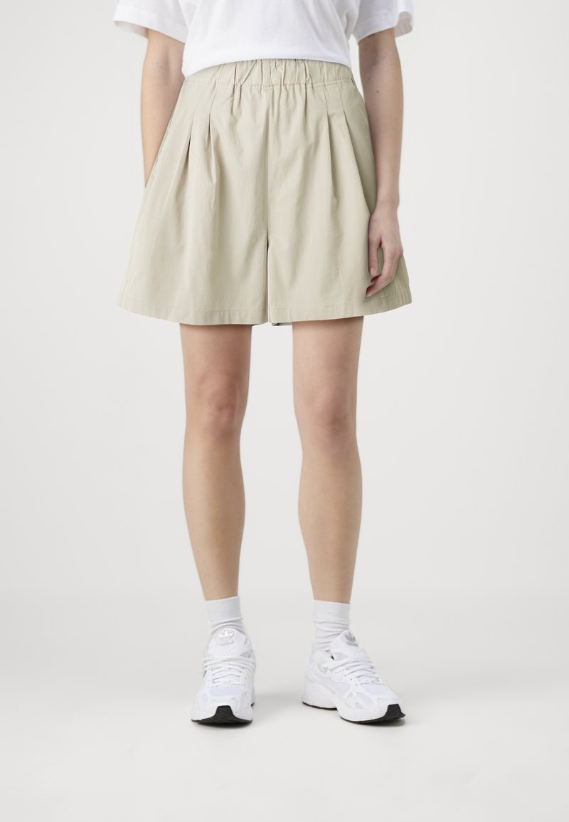 Shorts beiges plissés avec une taille élastique, fabriqués en tissu léger. Associés à des chaussures de sport blanches et des chaussettes mi-hautes.