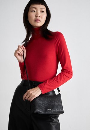 Jonge vrouw die een rood top met lange mouwen en een zwarte broek draagt, houdt een zwarte Calvin Klein schoudertas vast tegen een lichte achtergrond.