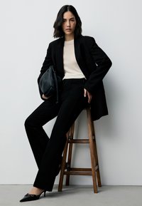 Black tailored blazer and high-waisted trousers, paired with a cream ribbed top. She carries a black leather bag and wears pointy heels.