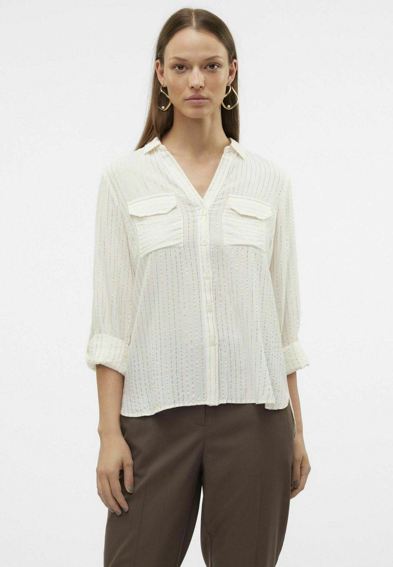 Blouse blanche à manches longues avec un devant à boutons, deux poches poitrine, des rayures verticales et des manches retroussées, associée à un pantalon marron.