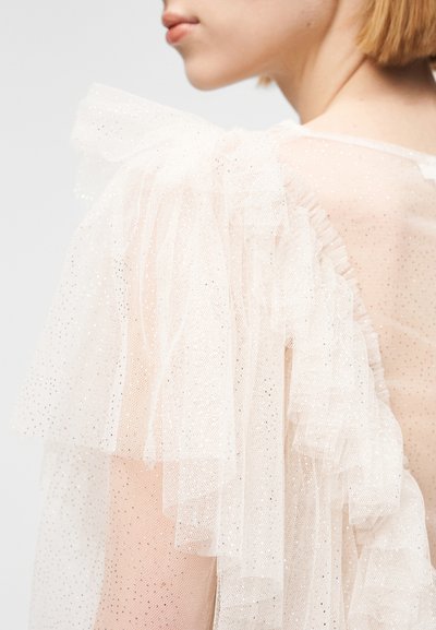 Αμάνικο φόρεμα με διαφανές τούλινο ύφασμα, ανοιχτό μπεζ χρώμα, διακοσμημένο με πινελιές από βολάν και διακριτική γκλίτερ για υφή και λεπτομέρεια.