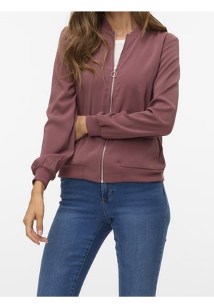 Femme portant une veste bomber mauve zippée avec des poignets côtelés et un jean bleu sur un fond blanc uni.