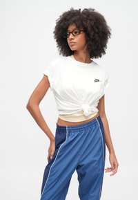 Λευκό βαμβακερό T-shirt με κόμπο μπροστά, με μικρό μαύρο λογότυπο Nike. Συνδυασμένο με μπλε άνετο αθλητικό παντελόνι με λευκές λεπτομέρειες.