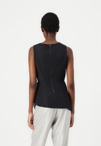 Svart ermeløs topp med peplum-design og glidelås i midten bak, laget av glatt stoff. Kombinert med lysegrå, glinsende vide bukser.