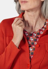 Rode gebreide cardigan met een kraagnek en twee rode knopen. Daaronder een top met patronen van geometrische vormen in rood, bruin en blauw.