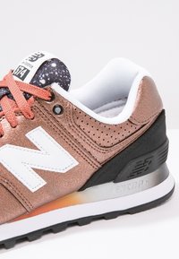 Gros plan d'une basket New Balance 574 bronze avec logo blanc, soutien de talon noir, lacets corail et languette noire et blanche tachetée.