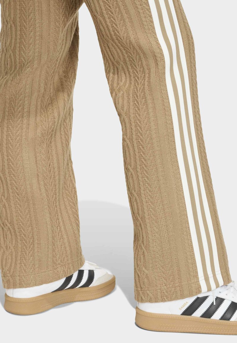 Beige cable-knit pants with flat, wide leg design, featuring white vertical stripes. Paired with white sneakers showing black stripes.