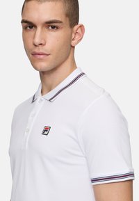 Vit polotröja i bomull med en tvåknappsknäppning och en Fila-logotyp på vänster bröst. Kragen har röda och marinblå ränder.