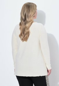 Langärmliges Creme-Oberteil mit rundem Ausschnitt, weicher Textur und lockerer Passform, kombiniert mit schwarzen Hosen, die ein Leo-Print-Detail aufweisen.