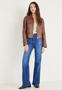 Barna bőr rövidített dzseki elöl cipzárral, világoskék trapéznadrággal párosítva. A modell pöttyös felsőt és fekete szegecses cipőt visel.