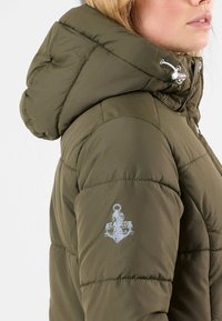 Olijfgroene gewatteerde jas met capuchon, voorzien van zilveren hardware en een geborduurd logo op de mouw. Gladde textuur en gequilt ontwerp.