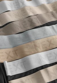 Elastické pásky v různých odstínech, včetně béžové, šedé a černé, s raženým logem "NEXT". Hladká textura, jednotná šířka.