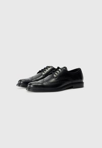 Chaussures de brogue en cuir noir avec un design à lacets, présentant des détails perforés et un bout arrondi. Semelle en caoutchouc plate offrant de la stabilité.