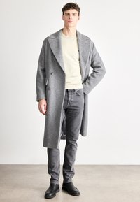 Γκρι παλτό από μείγμα μάλλινων υφασμάτων με σχέδιο ψαροκόκκαλο, διπλό στήθος, μεγάλο γιακά, φορεμένο πάνω από μπεζ πουκάμισο και σκούρο γκρι τζιν, με μαύρα παπούτσια.