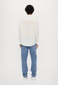 Bela, dolga srajca z dolgimi rokavi, narejena iz lahke tkanine, ima klasičen ovratnik in gumbaste manšete, v kombinaciji z sproščenimi modrimi denim hlačami.
