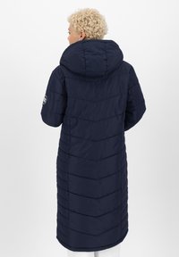 Marineblauwe gewatteerde jas met een grote capuchon, voorzien van een chevron-quiltpatroon en een logo-detail op de linker mouw. Lange uitvoering, gestructureerde stof.