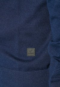 Pull en maille bleu foncé avec un tissu texturé et un petit patch en cuir noir au coin inférieur gauche.